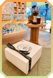 White gift box tied with a black diptyque ribbon on a green cloth, with a wooden table and colorful tiled wall in a bright shop interior.