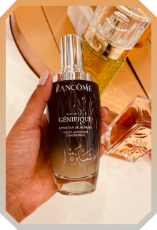 Hand holding a bottle of Lancôme Génifique youth activating concentrate serum with two other perfume or skincare bottles in the background.