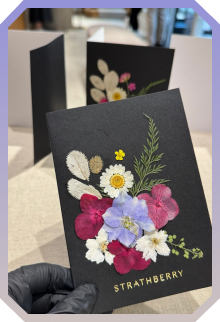 Black Strathberry card featuring colorful pressed flowers including daisies, pink, blue, and white blossoms with green foliage.