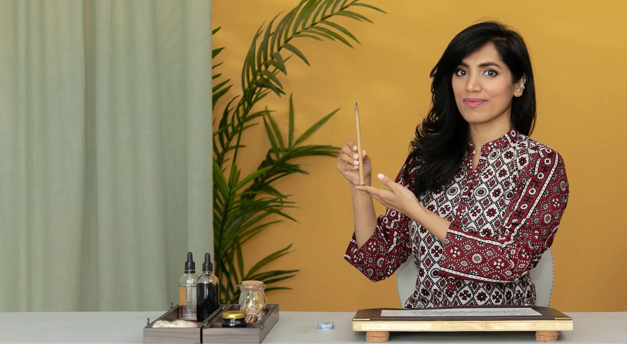 Woman with dark hair wearing a patterned top holding and presenting a wooden stick while sitting at a table with small bottles and a plant in the background.