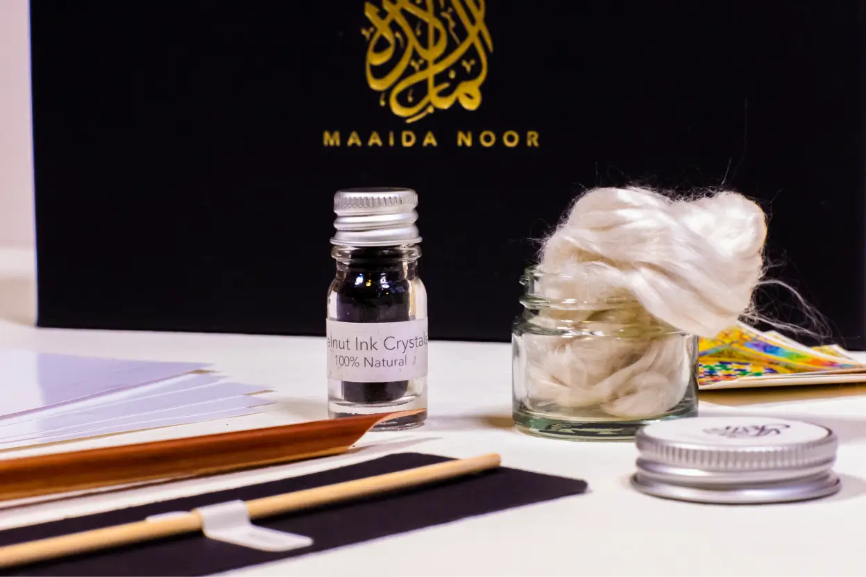 Calligraphy kit including a small bottle labeled 'Walnut Ink Crystals 100% Natural,' a jar with white fibers, wooden pen holders, and a black box with gold Arabic calligraphy and text 'MAAIDA NOOR'.