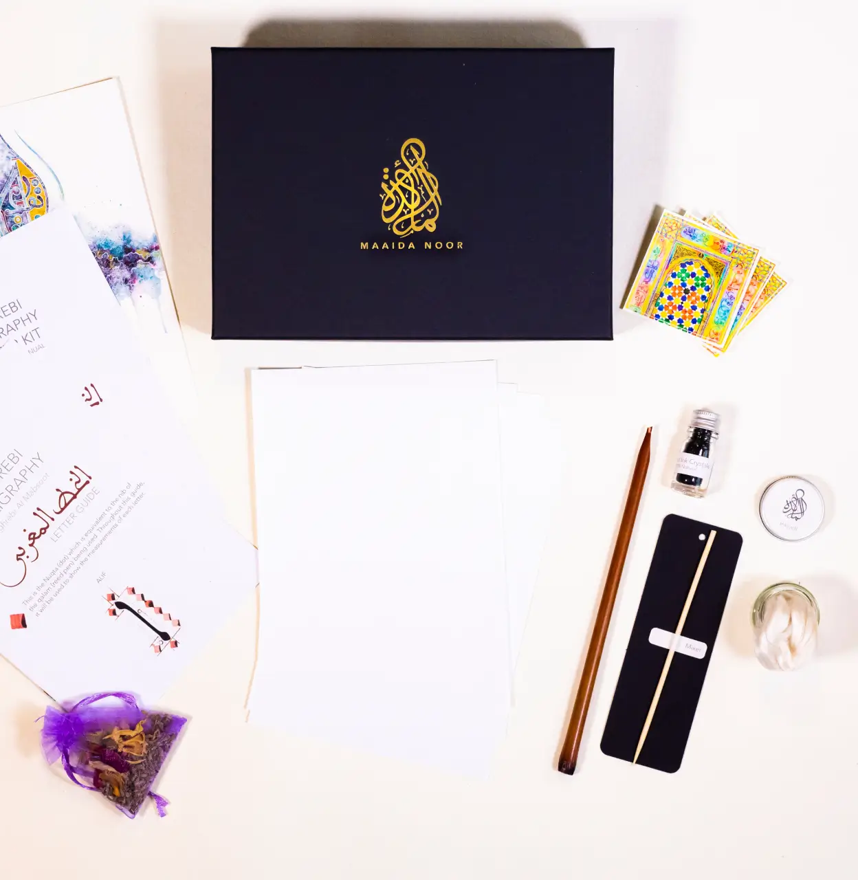 Calligraphy kit with black box, blank sheets, wooden pen, ink bottle, instruction booklets, decorative cards, cotton swabs, and dried flowers in a purple bag.