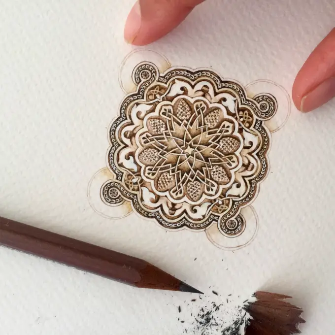 Close-up of a hand holding a detailed, circular mandala design drawn with a pencil on textured white paper, with pencil shavings nearby.
