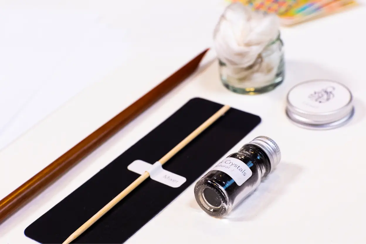 Calligraphy kit featuring a wooden pen, a stick labeled 'Mixer,' a small jar of black ink crystals, cotton pads in a glass jar, and a metal ink pot lid.