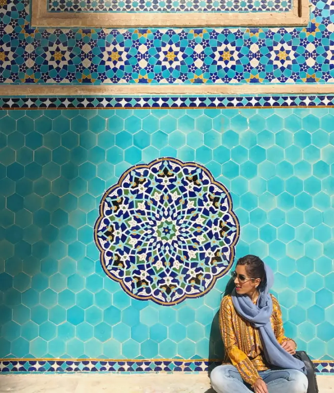Woman wearing sunglasses and a yellow patterned top sitting against a turquoise tile wall with a central intricate mosaic design.