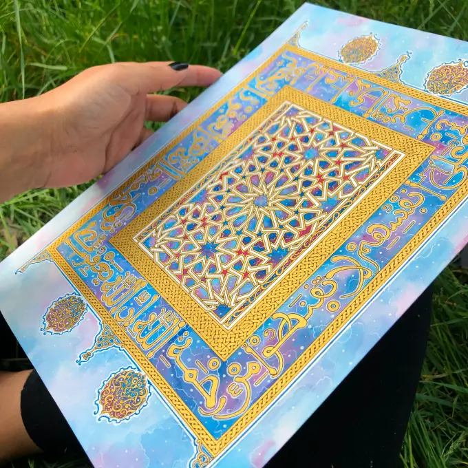 Hand holding a colorful Islamic geometric art piece with gold calligraphy on a blue and purple background, resting on grass.