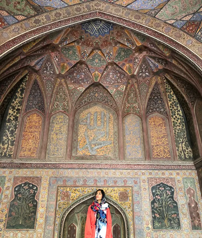 Woman wearing a colorful scarf and orange coat standing under a large ornate arch with intricate Islamic calligraphy and floral patterns.