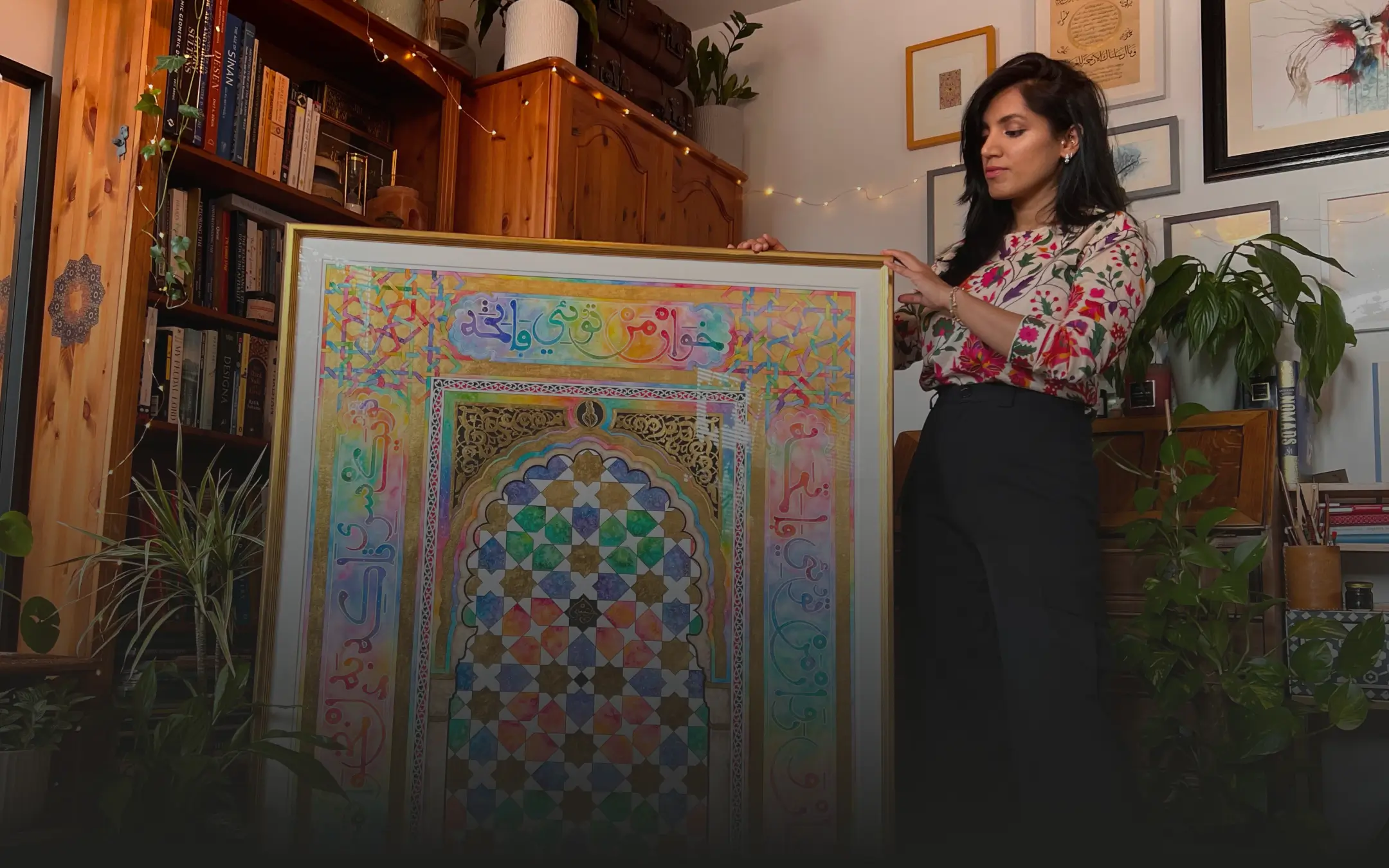 Woman holding a framed colorful geometric and calligraphy artwork in a cozy room with plants and wooden shelves.