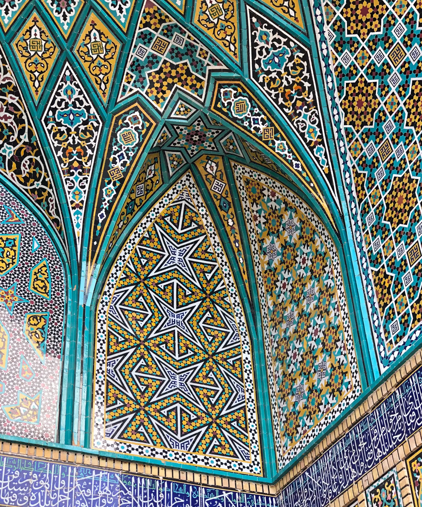 Intricate Islamic geometric and floral tile patterns in turquoise, yellow, white, and black decorating an arched wall and ceiling with Arabic calligraphy below.