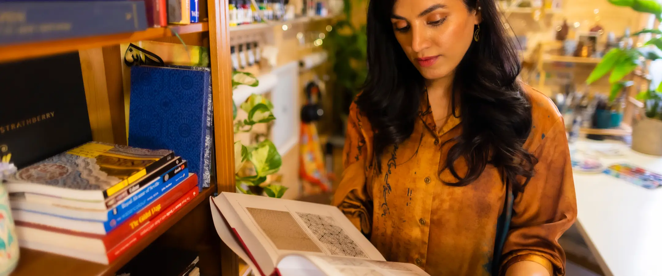 Woman in an orange shirt reading an open book next to a bookshelf with stacked books and plants.