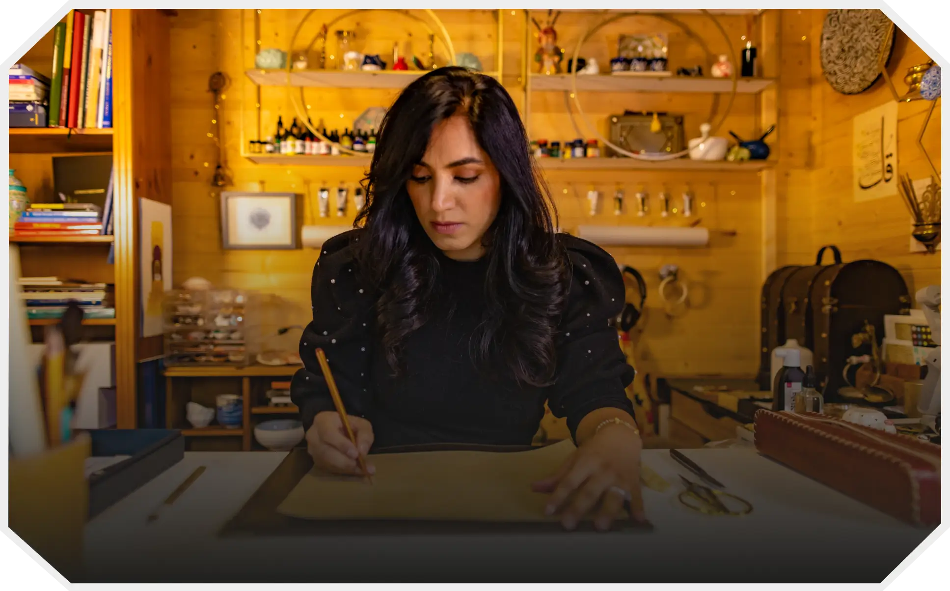 Woman with long dark hair focused on writing Arabic calligraphy script on paper at a cluttered wooden desk in a cozy, art-filled workspace.