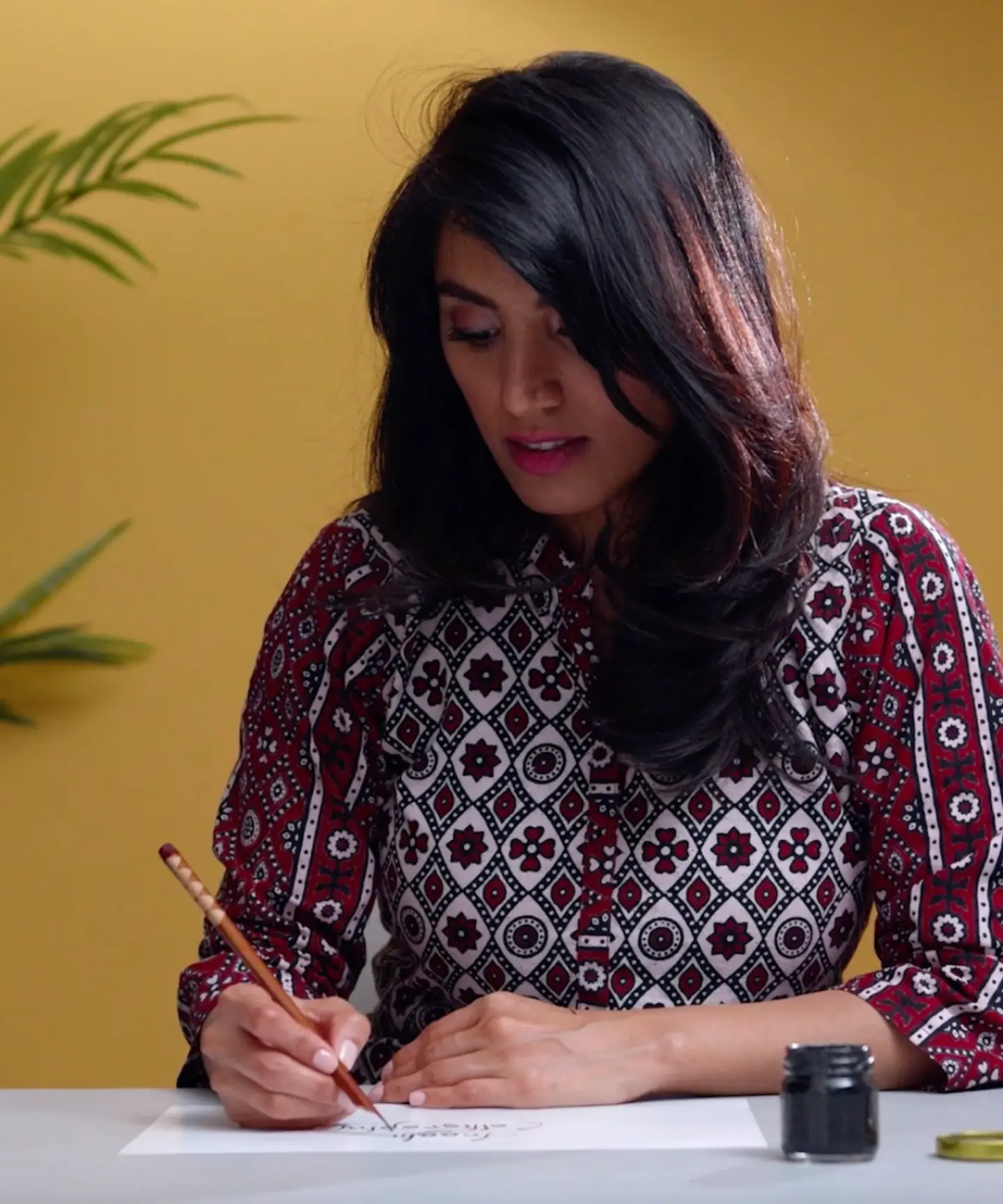Woman with dark hair wearing a patterned shirt writing calligraphy with a wooden pen and ink on white paper at a table.