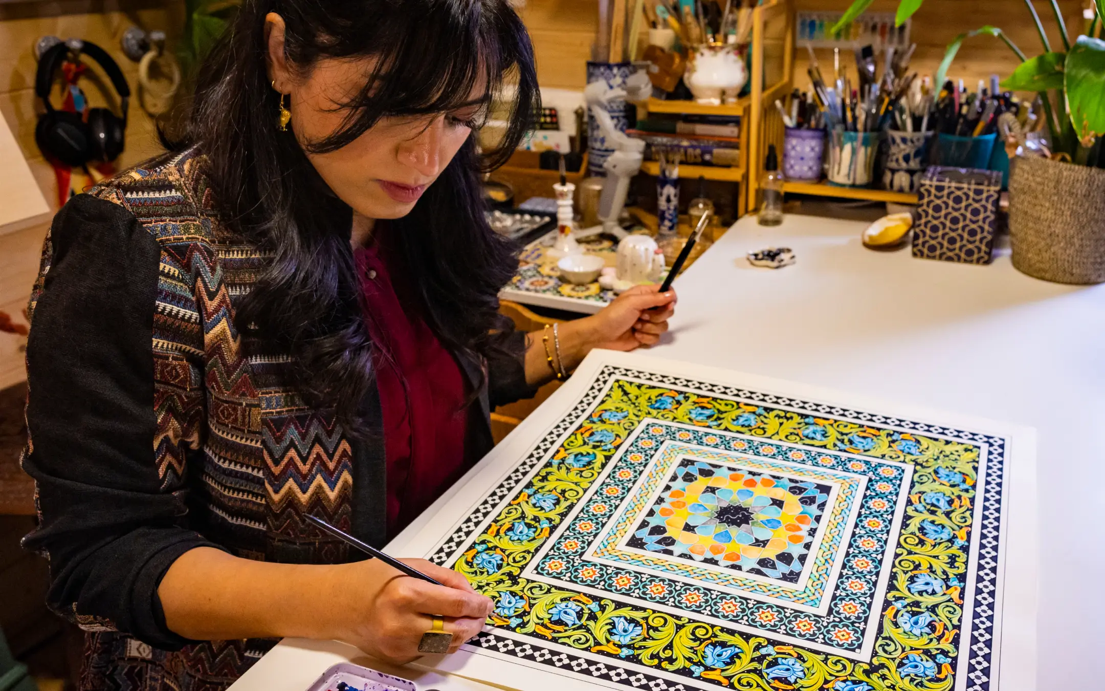 Woman painting an intricate geometric pattern with bright blue, yellow, and orange colors on a square surface at a desk.
