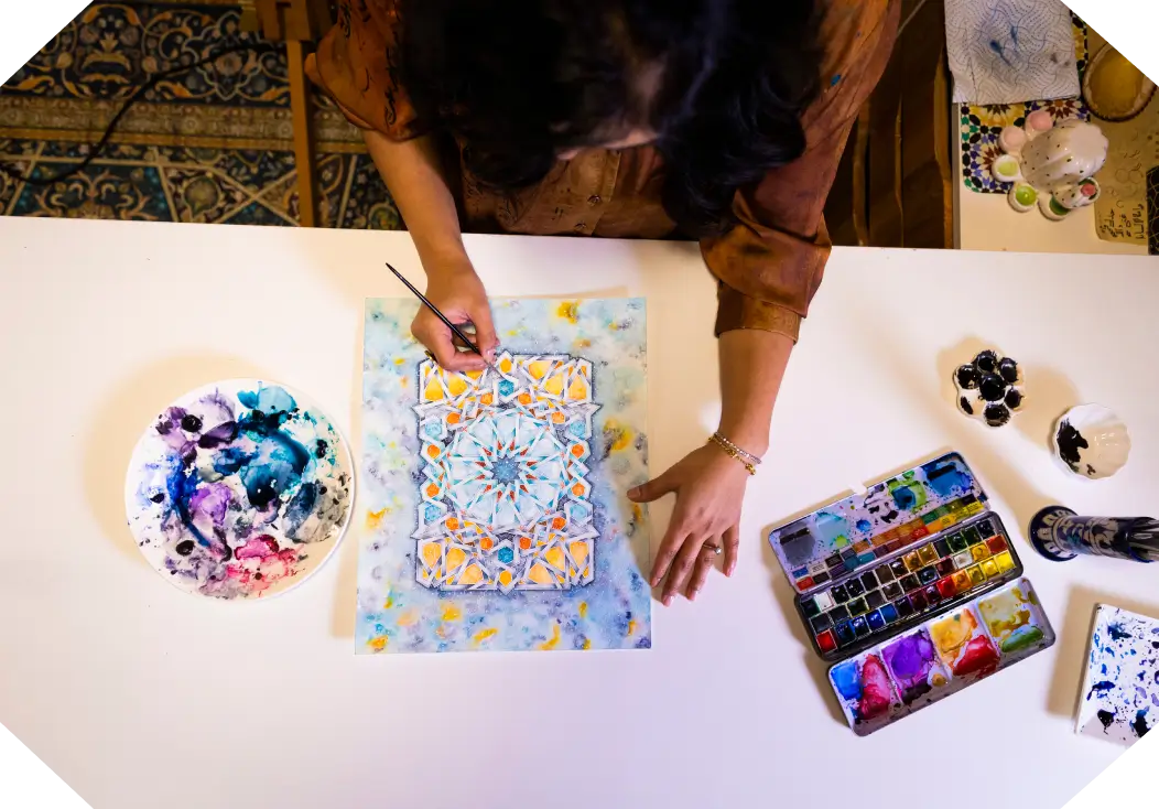 Top view of a person painting a colorful geometric mandala design on paper with watercolor paints on a white table.