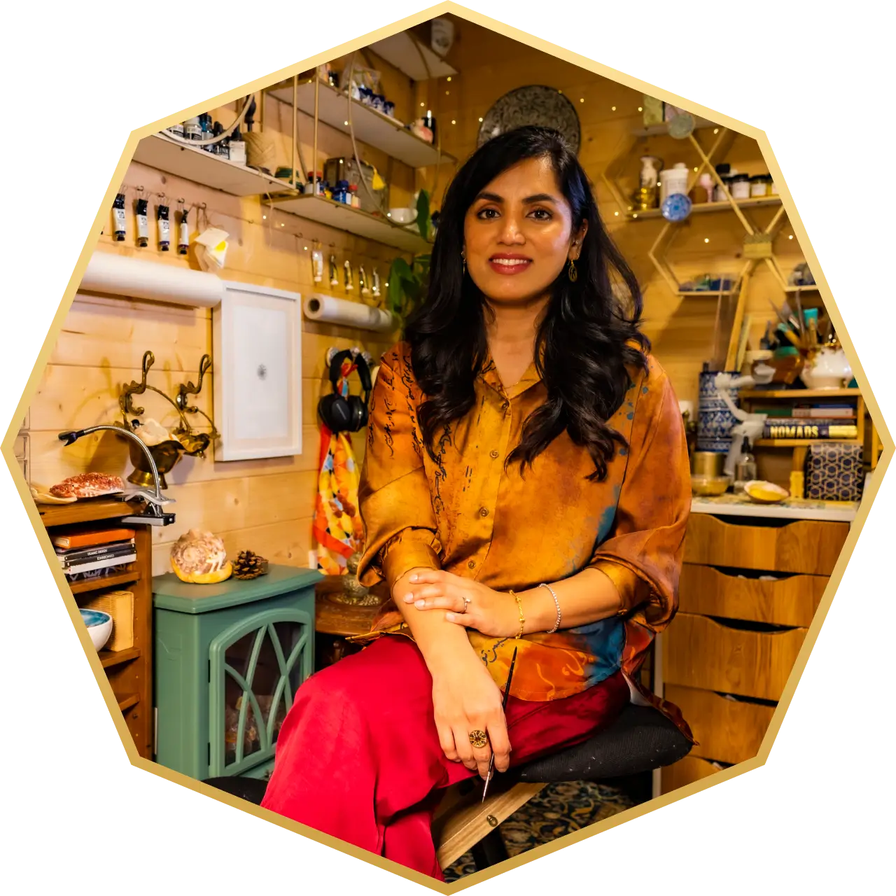 Smiling woman with long dark hair wearing an orange shirt and red pants sitting in a cozy, art-filled room.