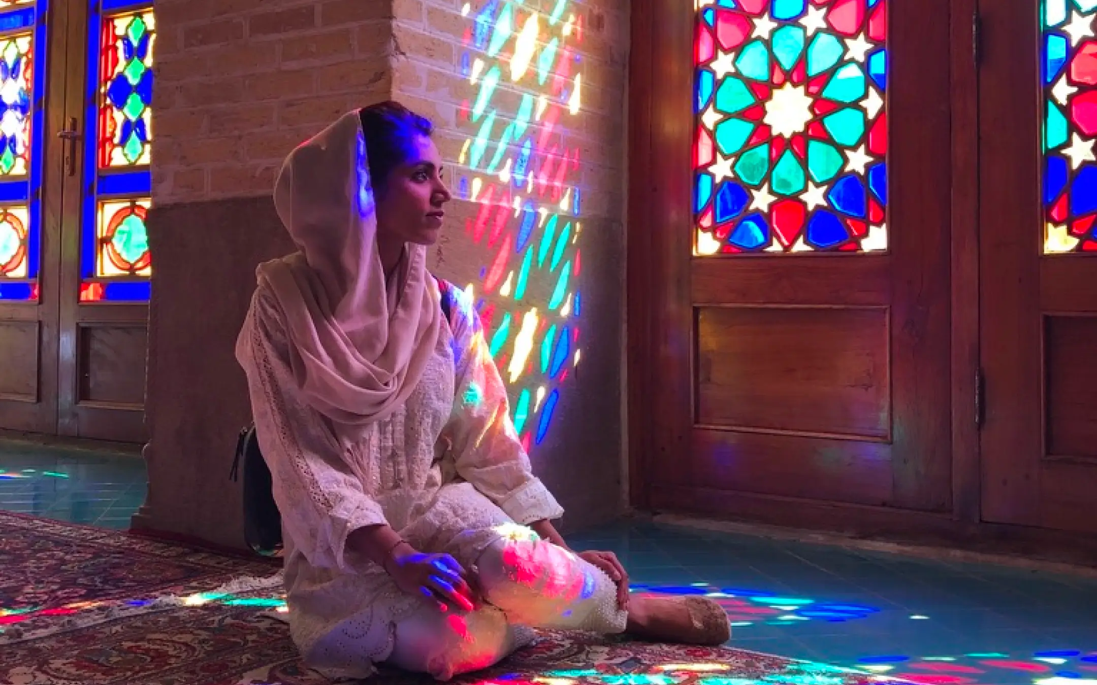 Woman in white traditional clothing sitting on a patterned carpet inside a building with colorful stained glass windows casting vibrant light patterns.
