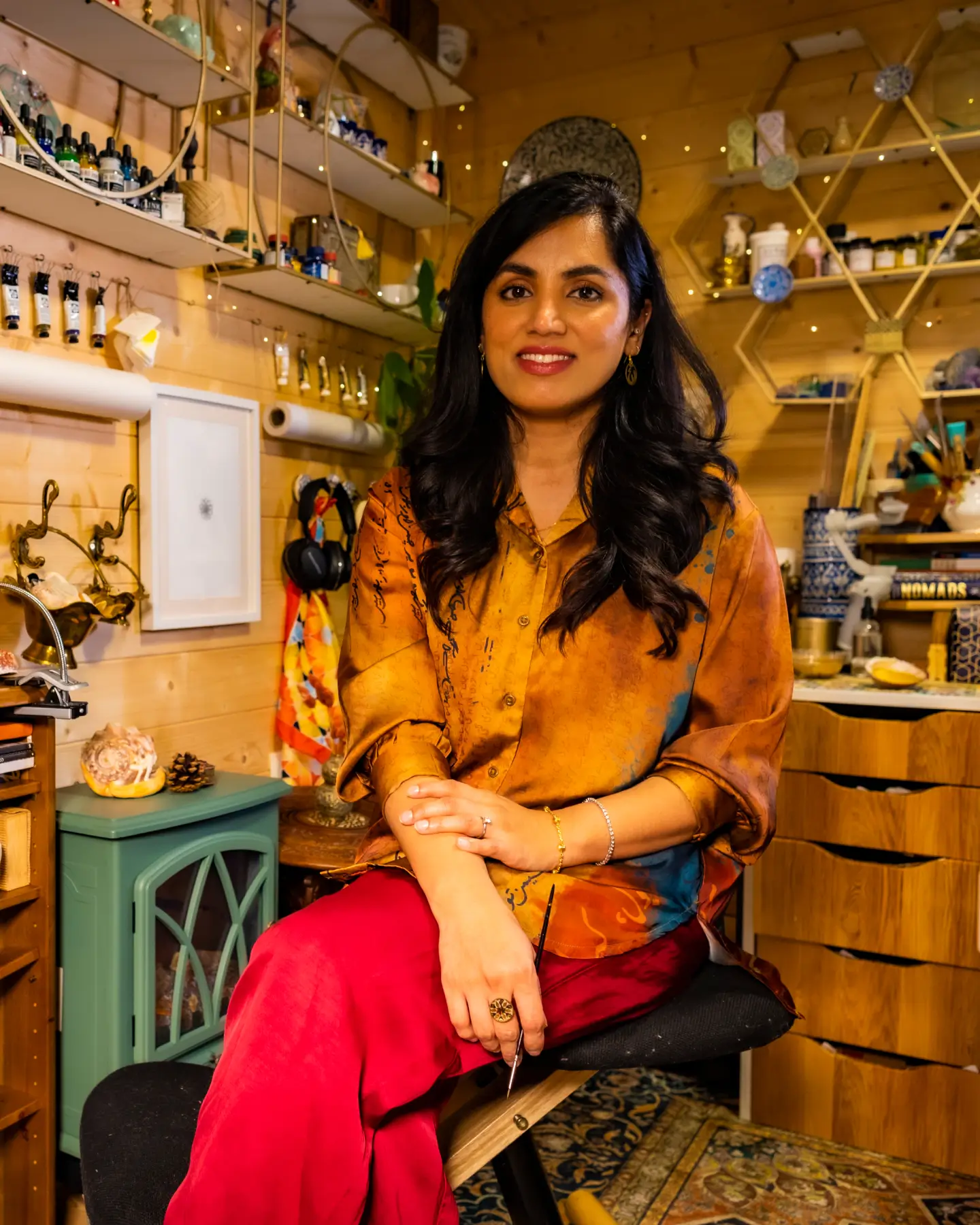 Smiling woman with long dark hair wearing an orange shirt and red pants seated in a cozy, art-filled room.