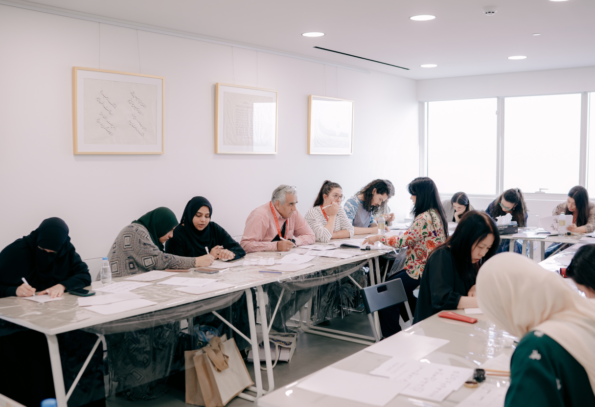 A diverse group of people seated at long tables in a bright classroom, engaged in Arabic calligraphy or drawing on paper.