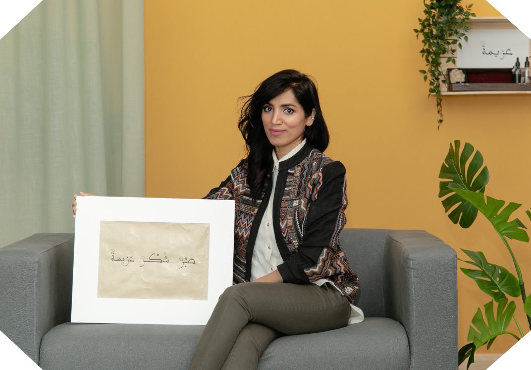 Woman sitting on a gray couch holding a framed artwork with Arabic calligraphy, with a yellow wall and green plants in the background.