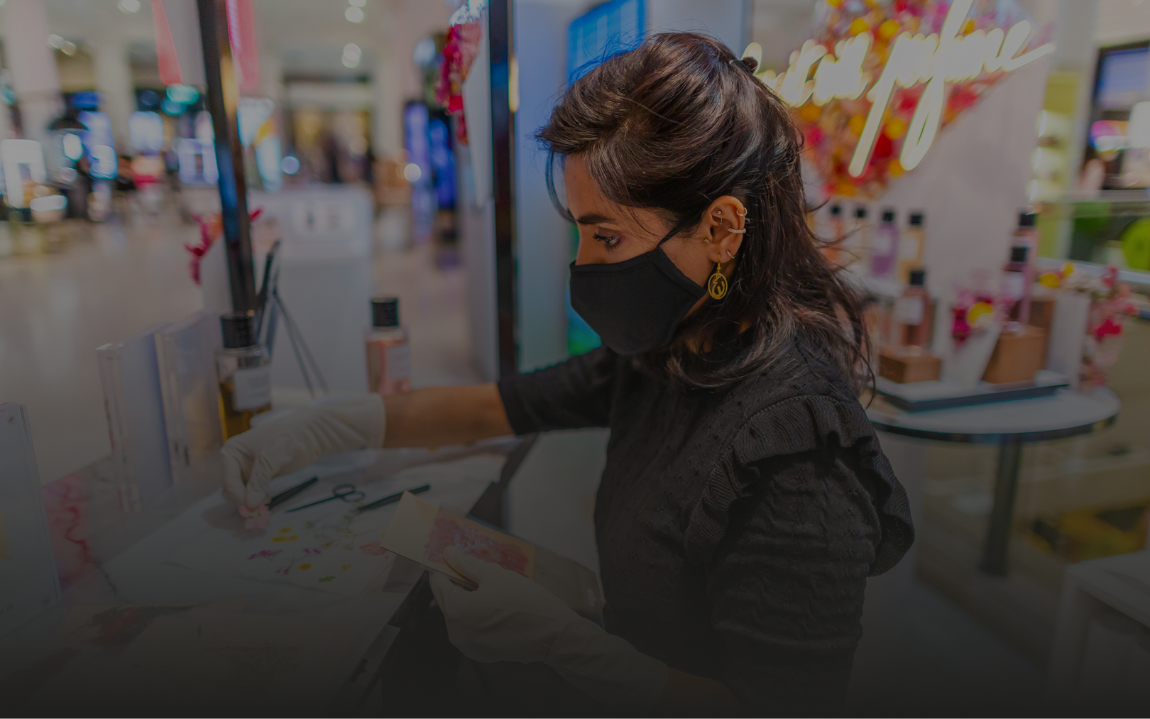 Woman wearing a black face mask and gloves working with colorful flower samples at a beauty or fragrance counter.