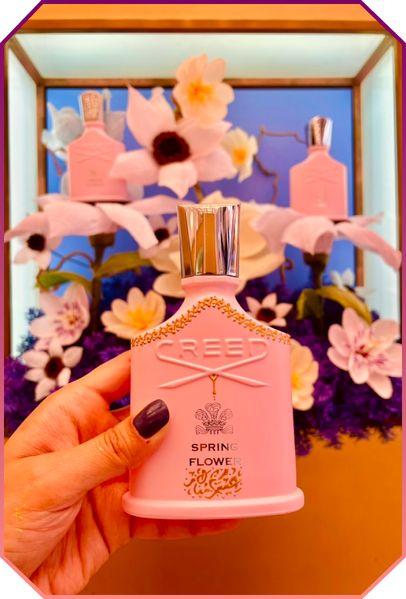 Hand holding a pink Creed Spring Flower perfume bottle with floral decorations in the background.