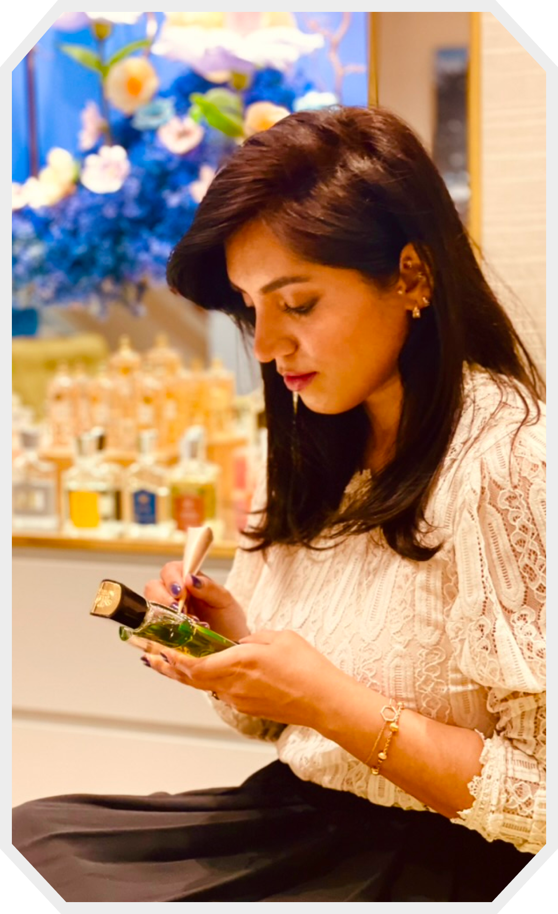 Woman artist doing calligraphy on a green perfume bottle indoors with a display of various perfumes and blue flowers in the background.