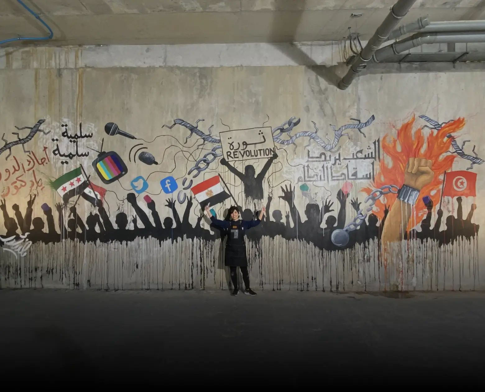 A person standing in front of a mural depicting a crowd with raised fists holding flags, social media icons, microphone, chains, and a sign reading 'Revolution' in Arabic and English.
