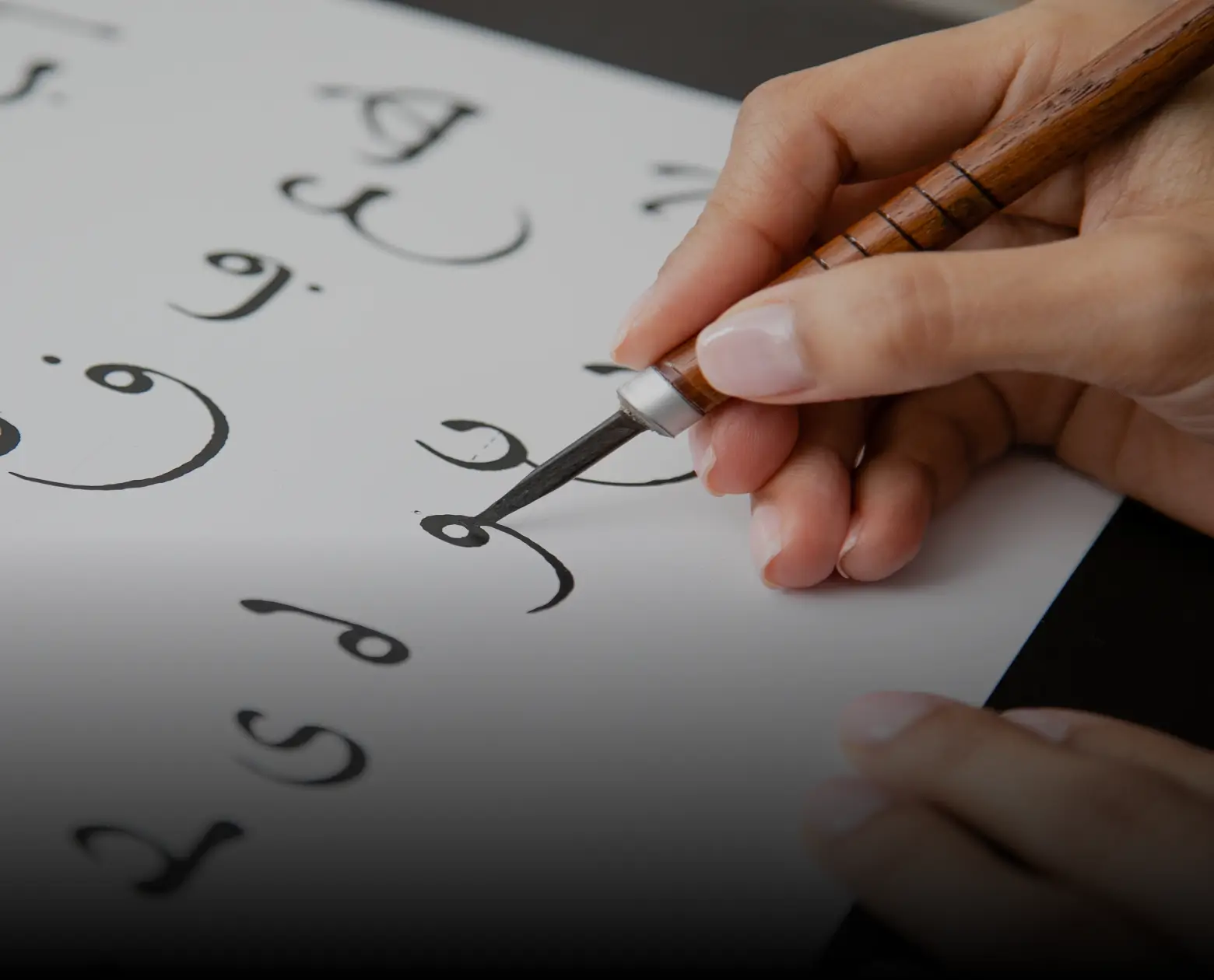 Hand holding a wooden calligraphy pen writing Arabic letters on white paper.
