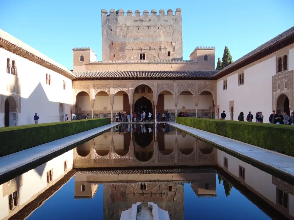 The Alhambra palace courtyard with a reflective rectangular pool and visitors walking around, under a clear blue sky.