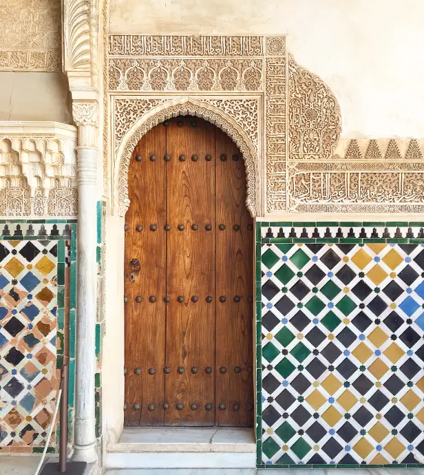 Ornate wooden door with arched frame surrounded by intricate Arabic-style plaster carvings and colorful geometric tiled walls.