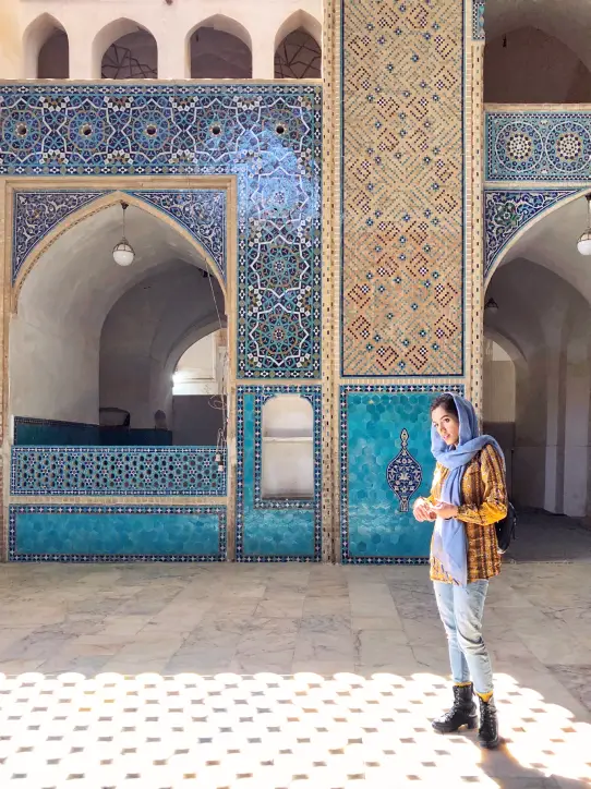 Woman wearing a blue headscarf and plaid shirt standing inside a building with intricate blue and beige tiled walls and arches.