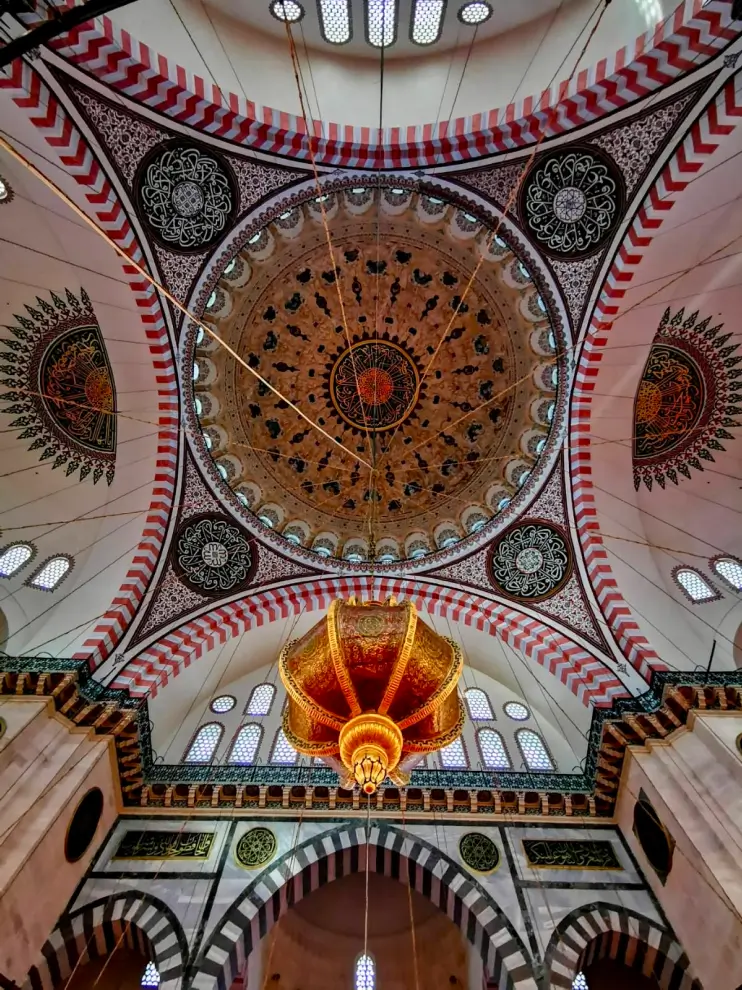 Intricately decorated mosque ceiling with a large central dome featuring geometric and floral patterns and a golden ornate chandelier hanging below.
