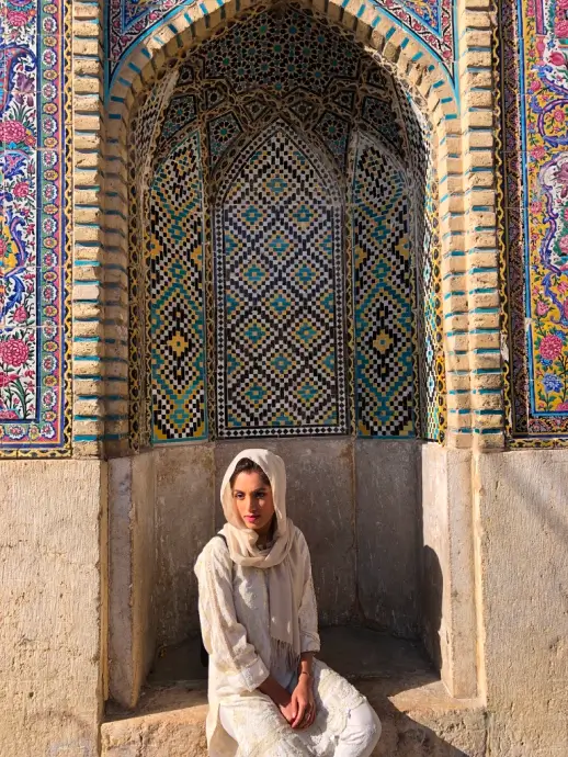 Woman in white traditional clothing sitting in front of an ornate tiled arch with geometric and floral patterns.