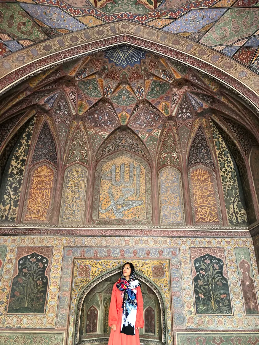 A woman in a red dress and colorful scarf stands under an ornately decorated arch with Islamic calligraphy and floral patterns on the walls and ceiling.