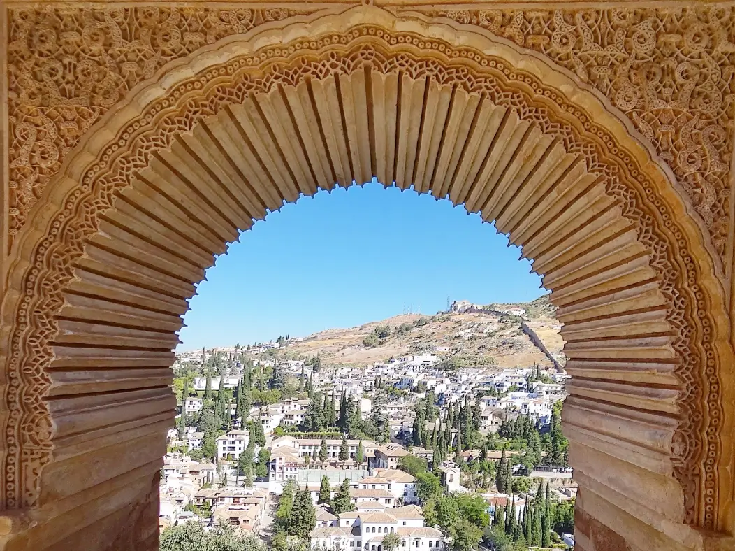 View of a hillside town with white buildings and green trees framed by an ornate stone arch with intricate carvings.