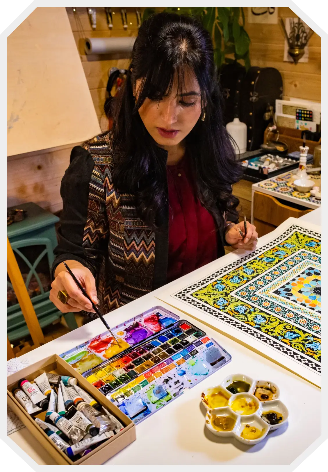 Woman painting a colorful intricate geometric pattern on paper with watercolor paints at a desk.