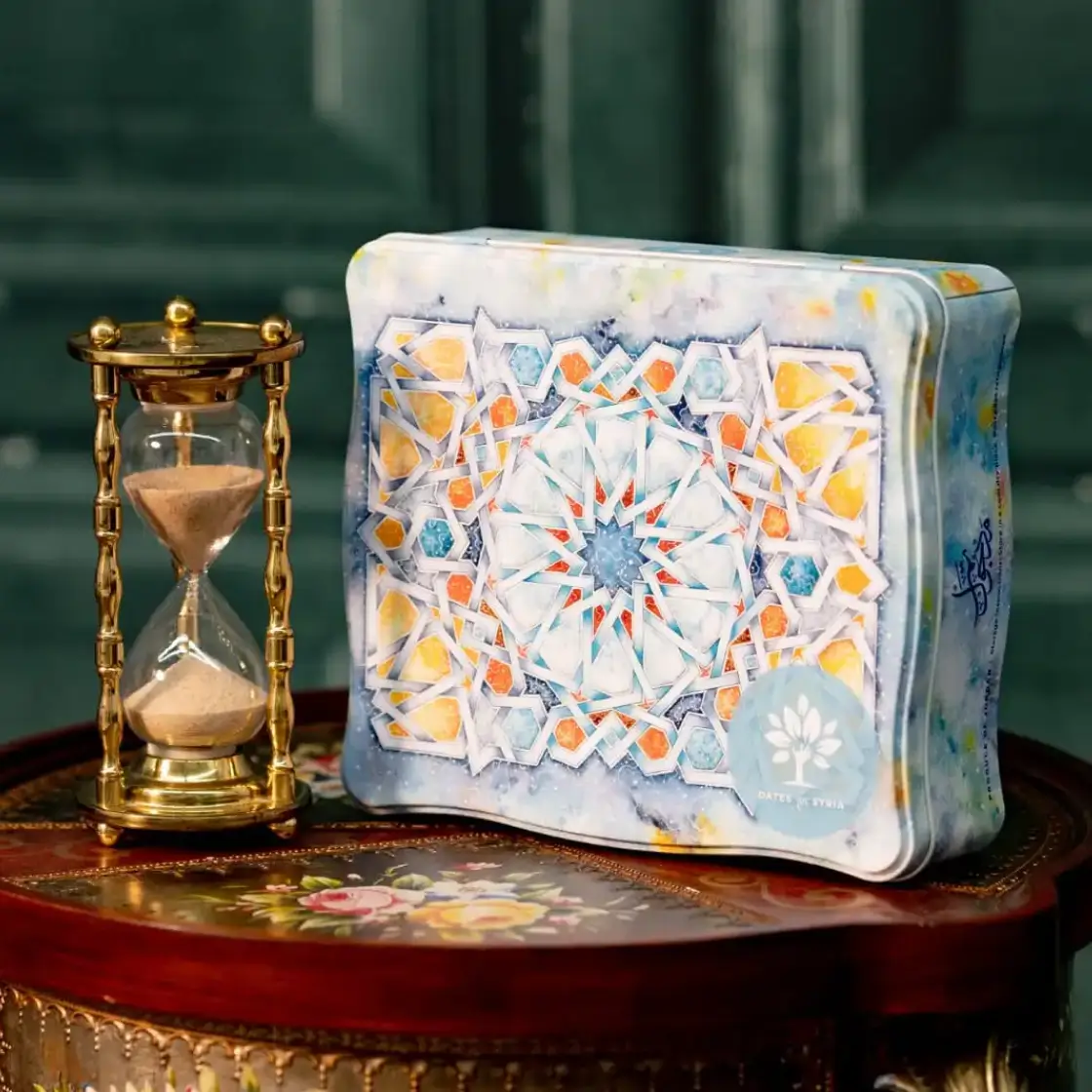 Ornate brass hourglass filled with sand beside a decorative tin box with colorful geometric patterns, placed on a wooden table with floral inlay.