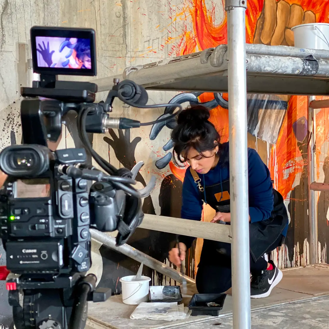A female artist painting a mural behind metal scaffolding while being recorded by a professional video camera.