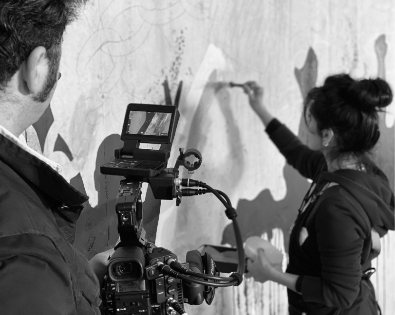 Black and white shot of an artist painting a mural being filmed by a professional camera.