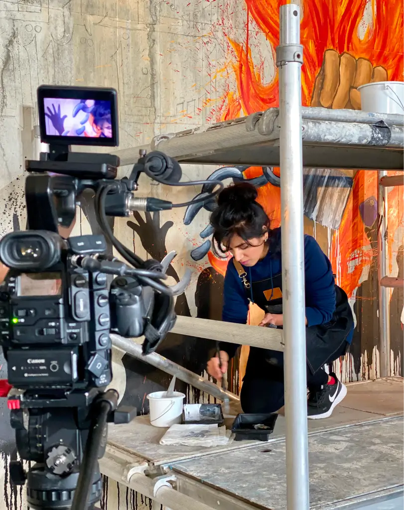 View through a film camera monitor of an artist working on a vibrant orange mural.