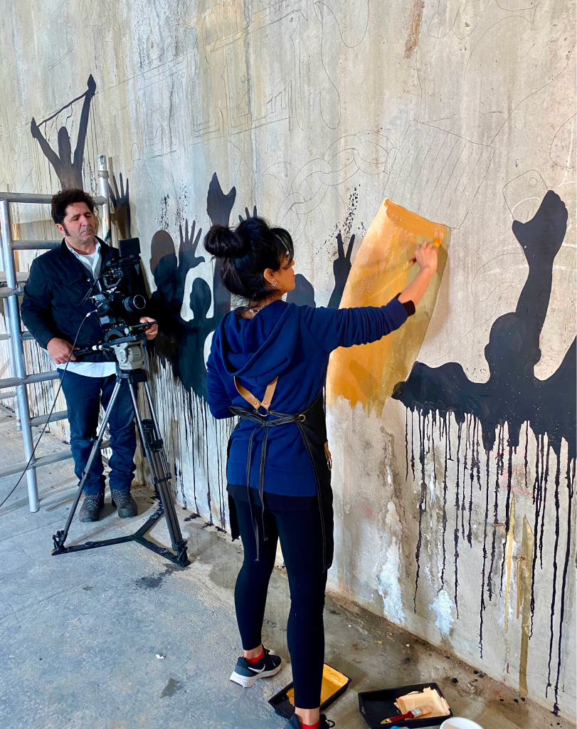 Artist applying a stencil to a wall mural while being recorded by a camera operator.