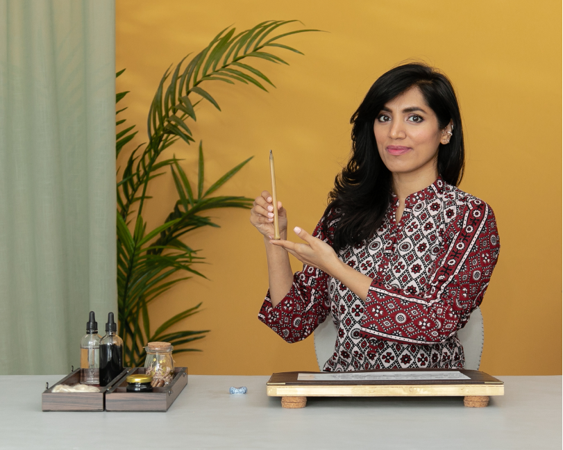 A female instructor in a patterned blouse holding a calligraphy pen (qalam) while demonstrating a technique in front of a yellow background with a houseplant.