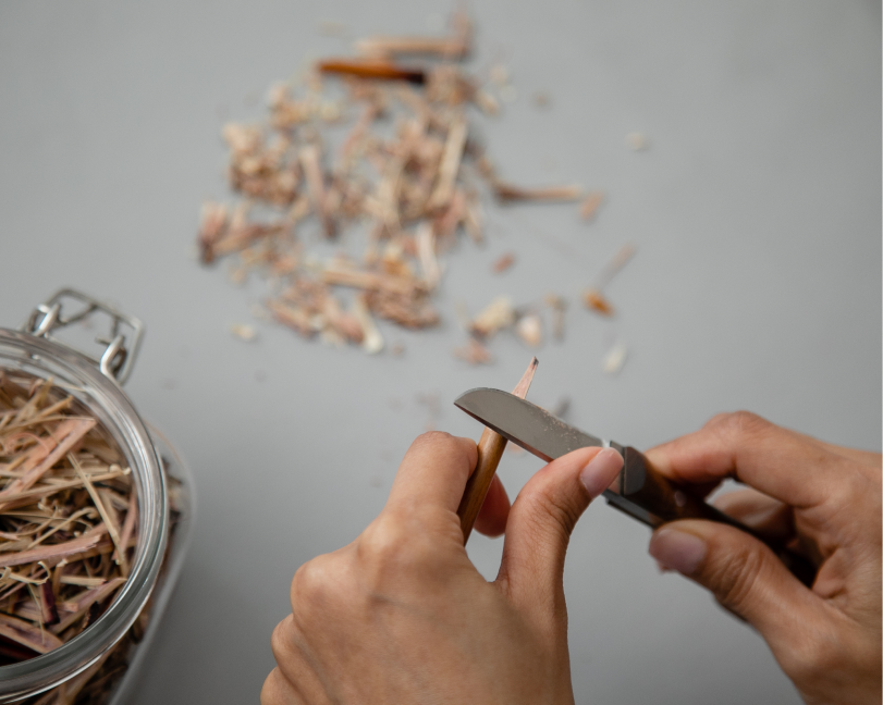 Close-up of hands using a craft knife to sharpen the tip of a bamboo or reed calligraphy pen, with wood shavings visible on the table.