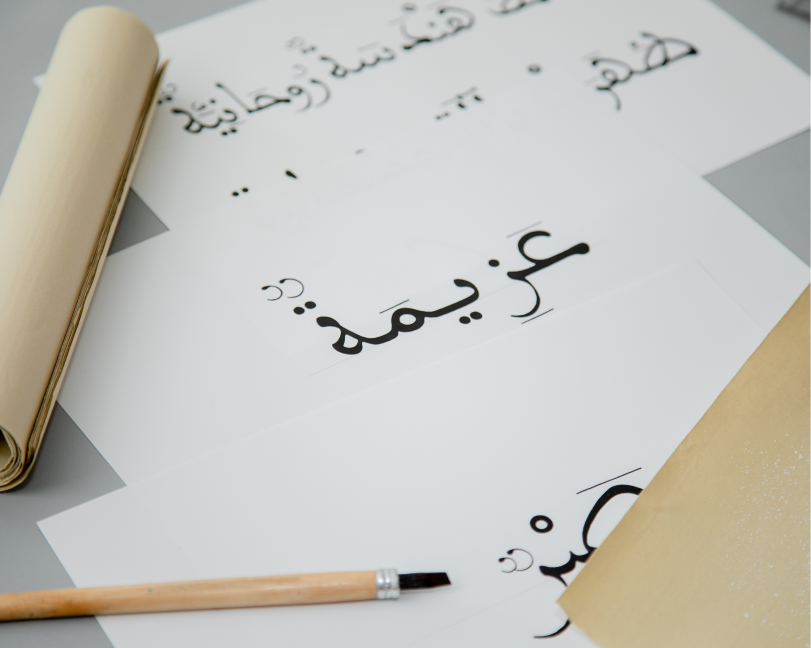 A wide-angle view of a calligraphy workspace showing a large Arabic word written on paper next to a rolled parchment and a reed pen.