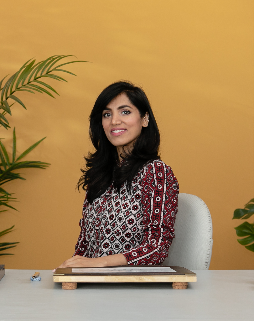 Portrait of a smiling female calligraphy artist sitting at her desk with her tools, positioned against a warm yellow backdrop.