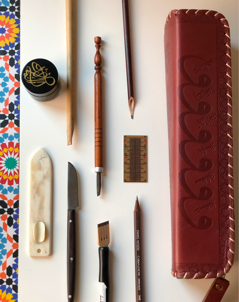 Overhead shot of a calligraphy toolkit including reed pens, ink wells, a leather pen case, and carving tools on a white surface.