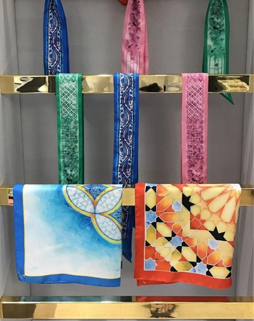 Collection of colorful patterned silk scarves hanging on a gold display rack, showing various blue, green, and orange cultural designs.