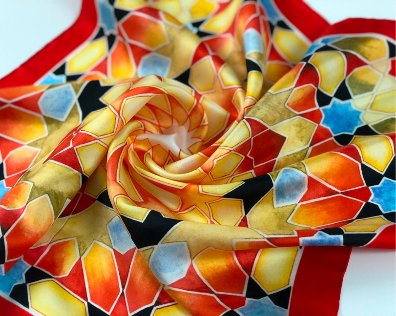 Stylized overhead shot of a silk scarf swirled into a rose shape, highlighting vibrant orange, red, and yellow mosaic-style geometric patterns.