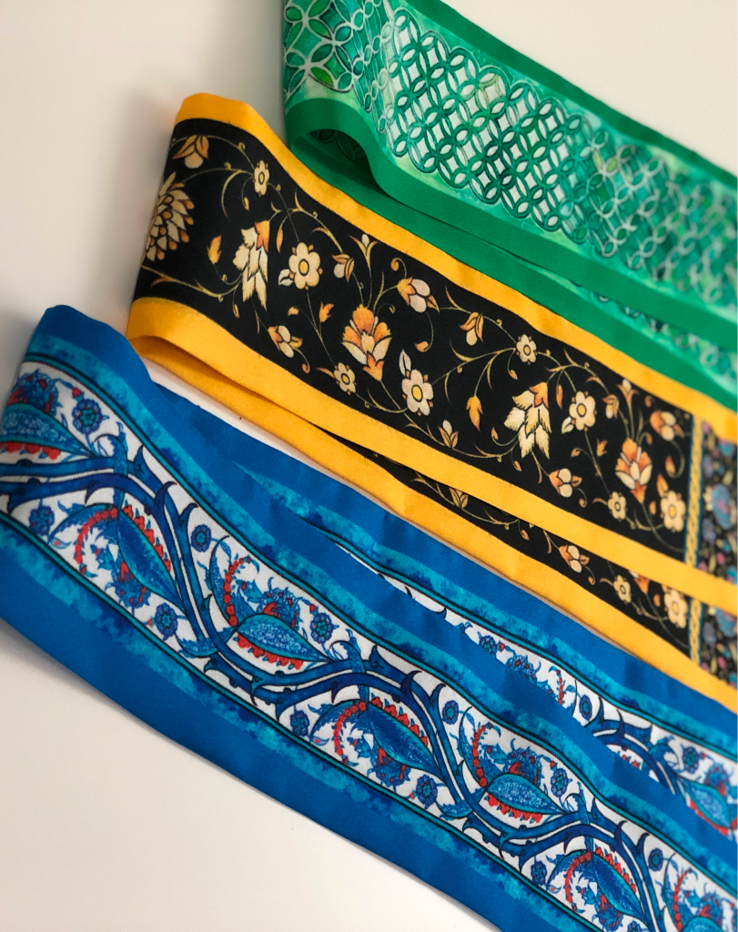 Three folded silk scarves in blue, black/gold, and green, showcasing diverse traditional Middle Eastern floral and geometric patterns.