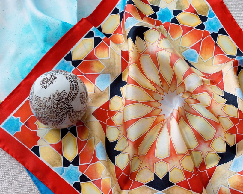 Close-up of a folded silk scarf featuring warm orange and red geometric Islamic star patterns with a light blue border.